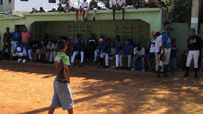 A Dominican child checks out the scene where tryout candidates await their turn in the dugout and neighborhood children pile onto the dugout roof.